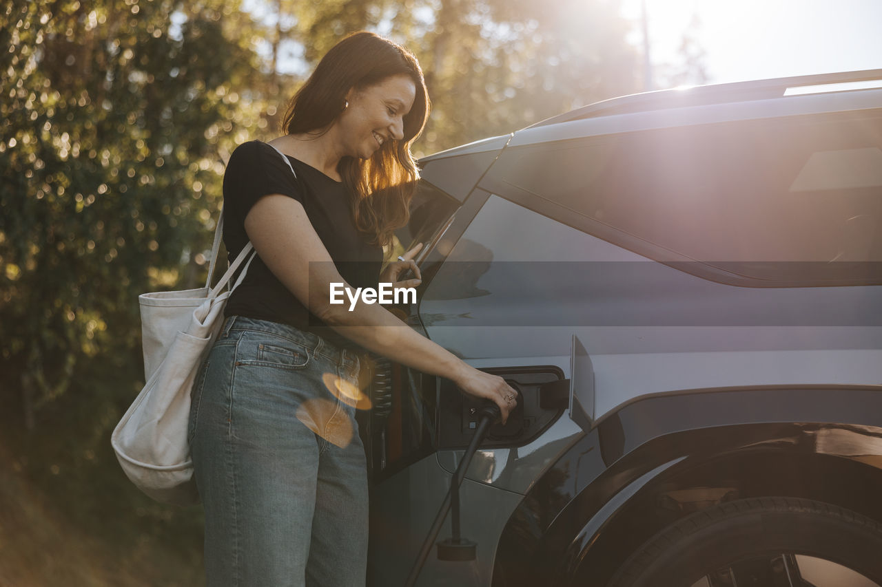Smiling mature woman charging car while standing at station