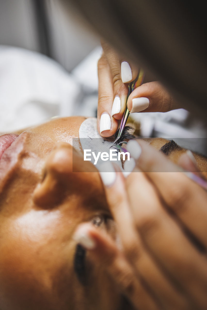 High angle of crop unrecognizable cosmetologist with tweezers applying fake eyelashes for extension on eye of ethnic client in salon