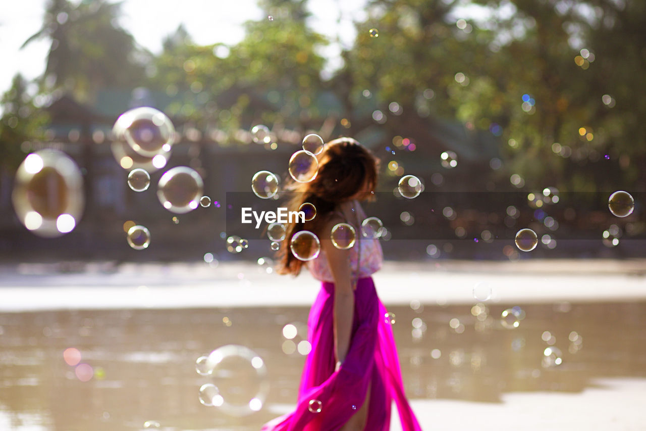 Side view of woman surrounded with bubbles while standing by pond