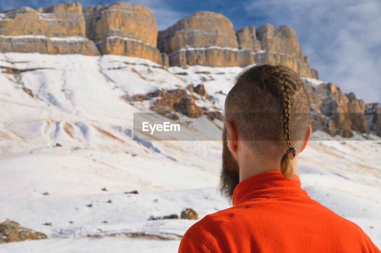 A bearded man with a stylish haircut in a red sweater stands with his back and looks into mountains