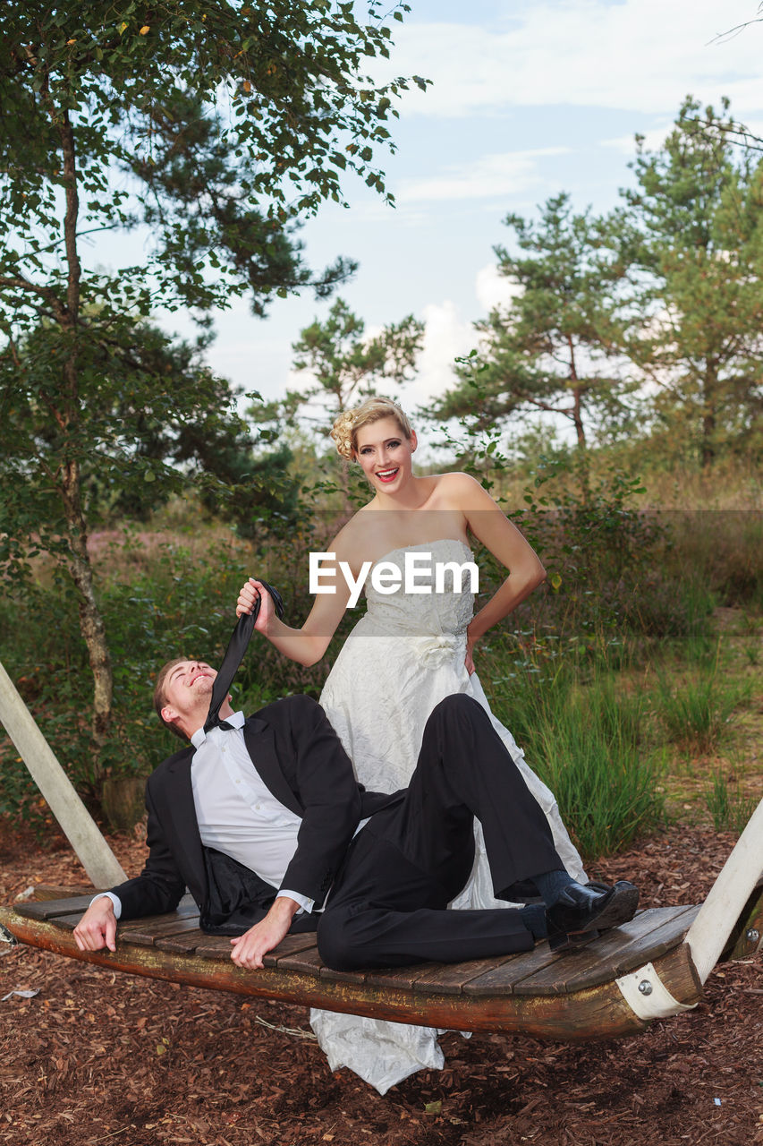 Portrait of playful bride holding necktie of bridegroom lying on swing at field