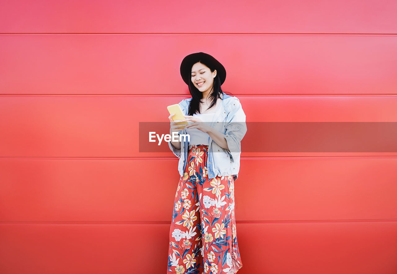 Smiling young woman using mobile phone against wall