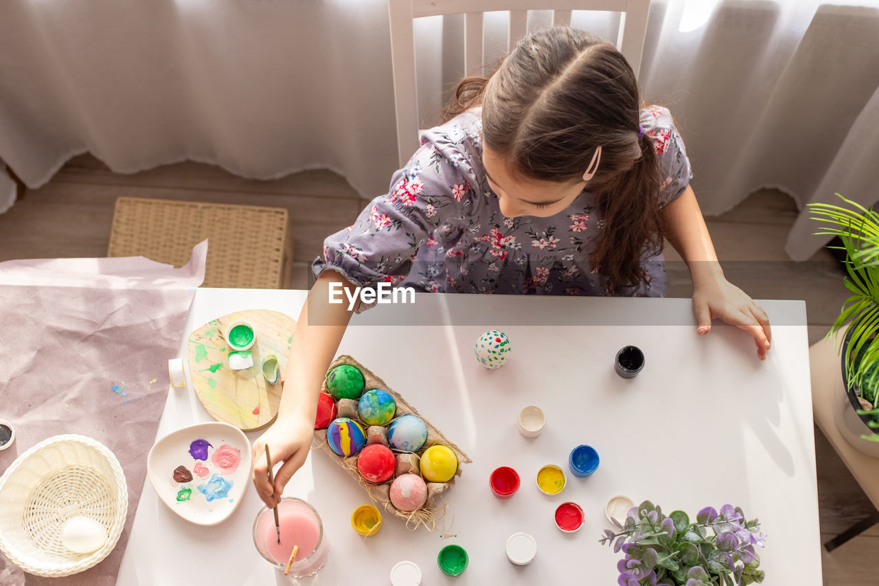 A cute little girl sits at a white table near the window, paints eggs with a brush.