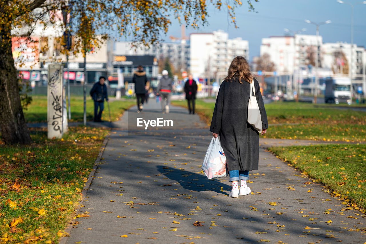 autumn, city, plant, spring, tree, nature, women, full length, adult, architecture, walking, road, one person, street, clothing, day, person, yellow, lifestyles, footpath, outdoors, rear view, focus on foreground, flower, city life, footwear, park, bag, leaf, park - man made space, young adult, female, building exterior, leisure activity, plant part, built structure, sunlight, casual clothing