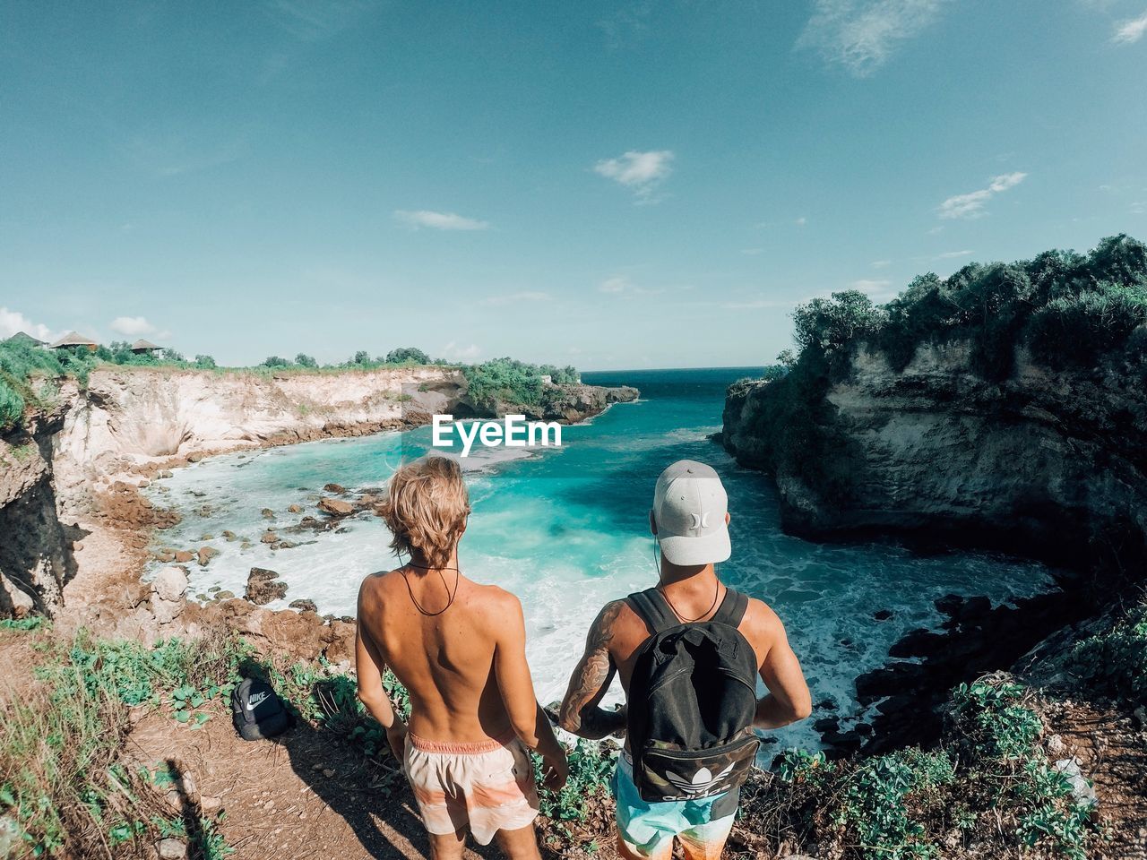 REAR VIEW OF FRIENDS SITTING ON ROCKS BY SEA