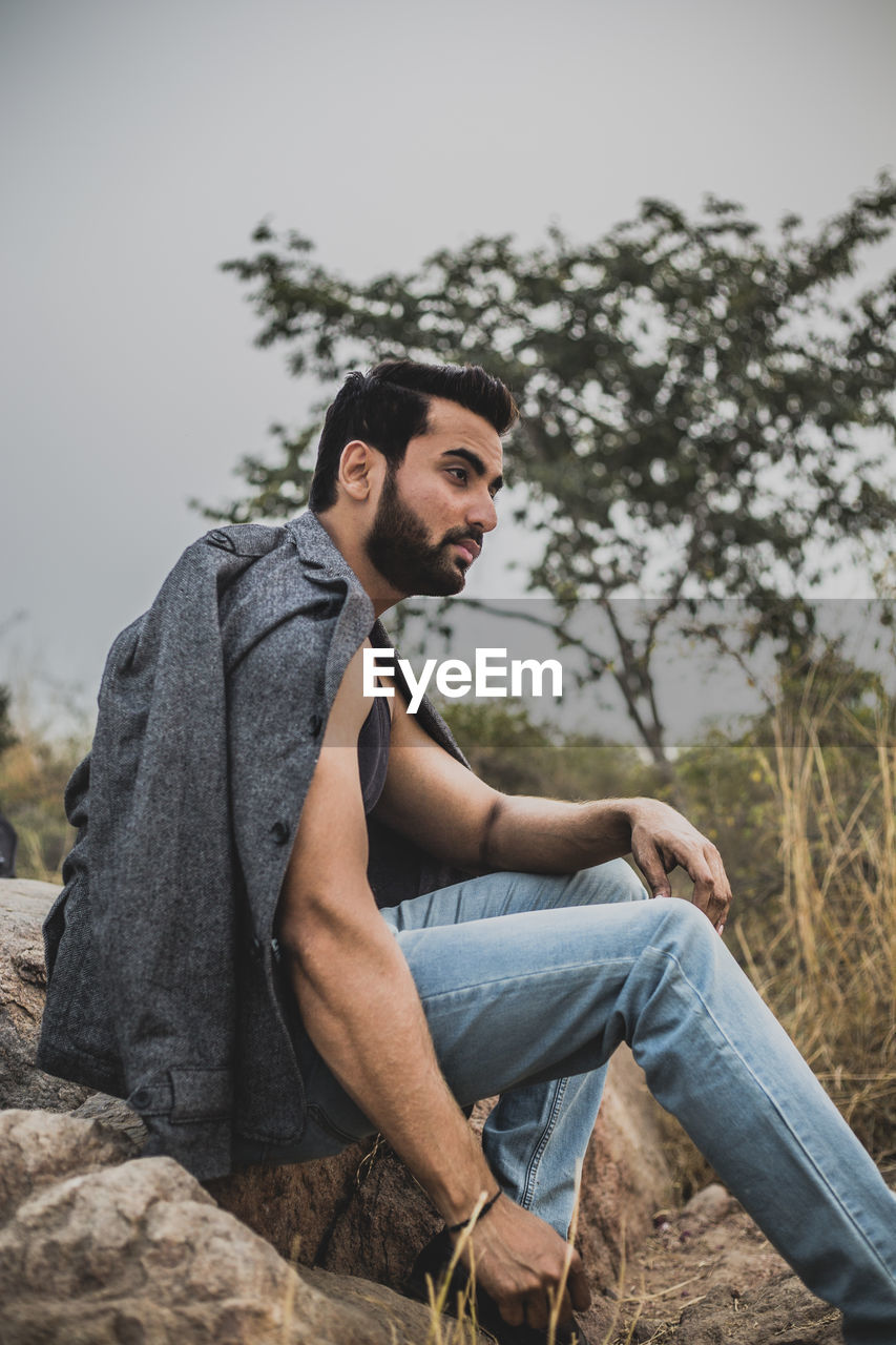 Side view of thoughtful young man sitting on rock against tree