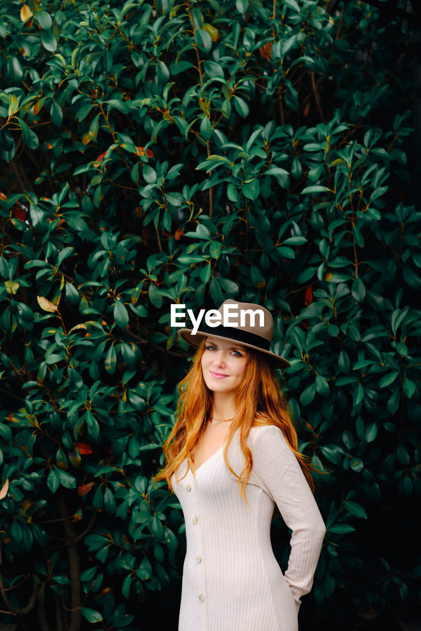 Attractive young long haired ginger female in stylish hat looking at camera while standing against green bushes in summer garden