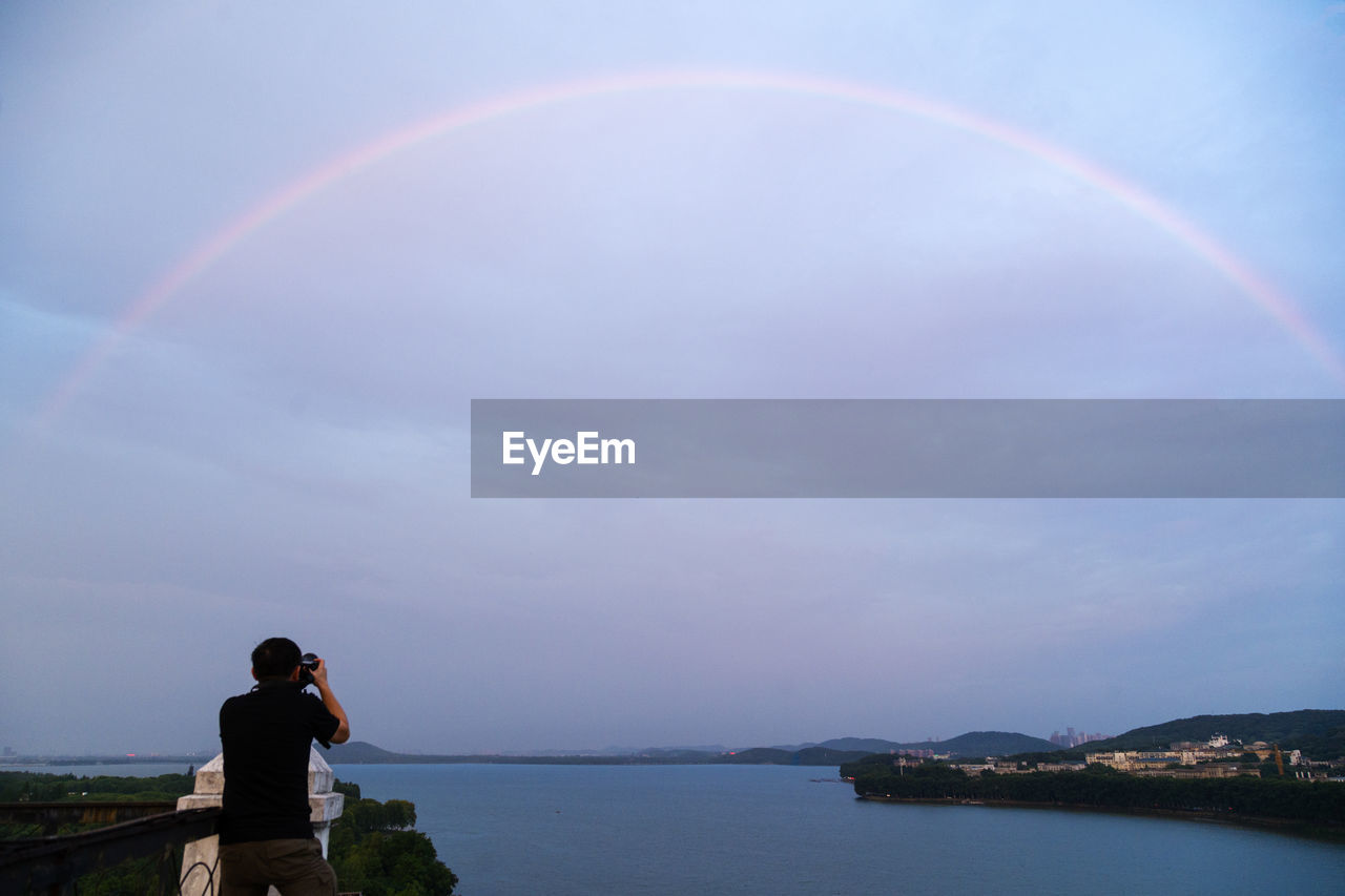 Rear view of man photographing rainbow over sea against sky