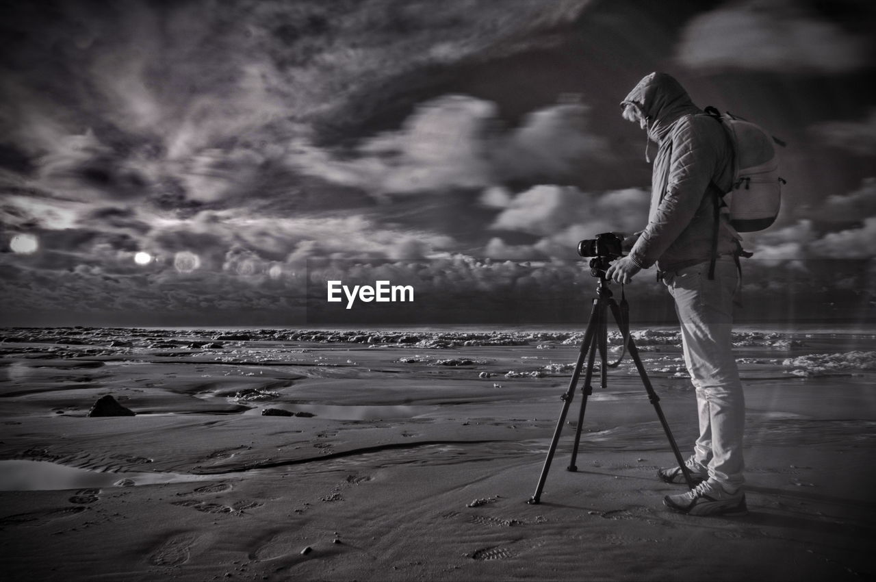 Side view of man with camera and tripod photographing at beach against cloudy sky