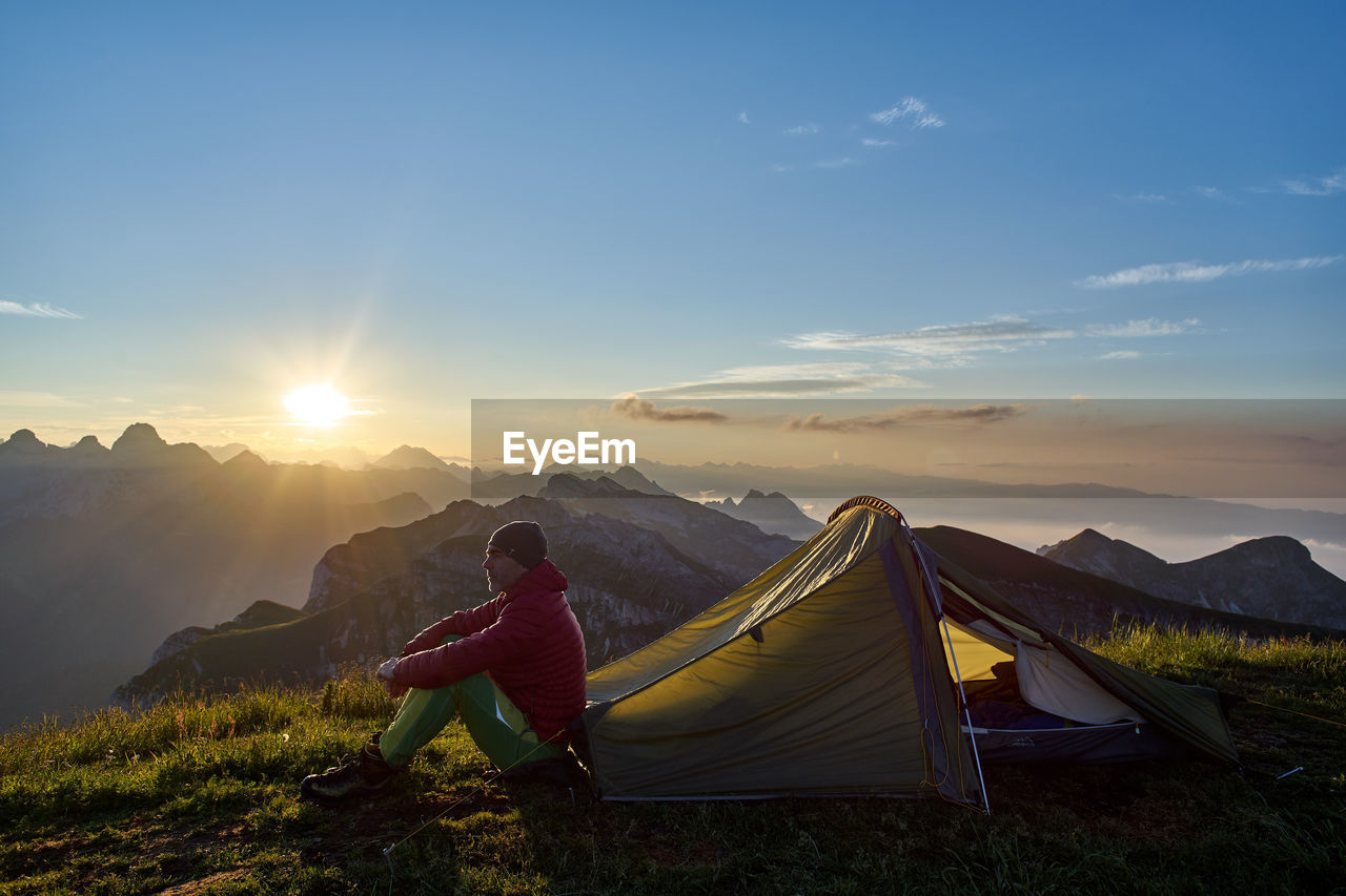 Full length of man looking at view while sitting on mountain by tent against sky during sunset