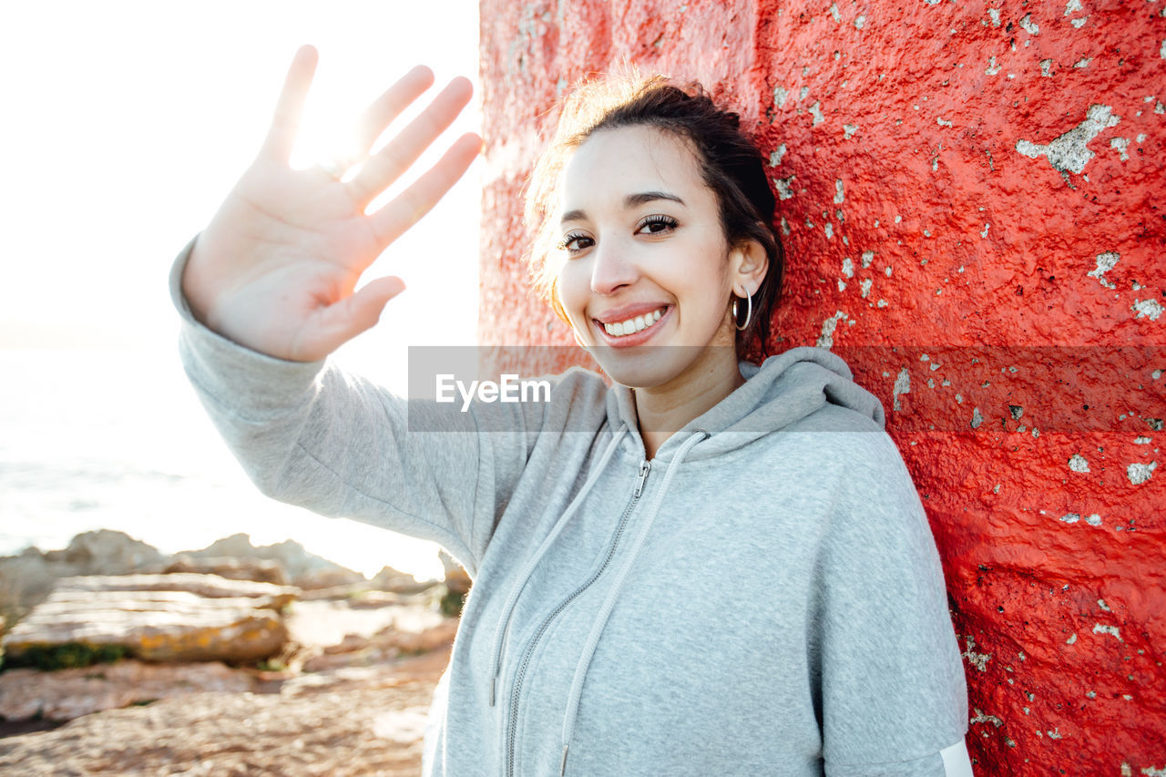 Portrait of young woman with arms raised standing against wall