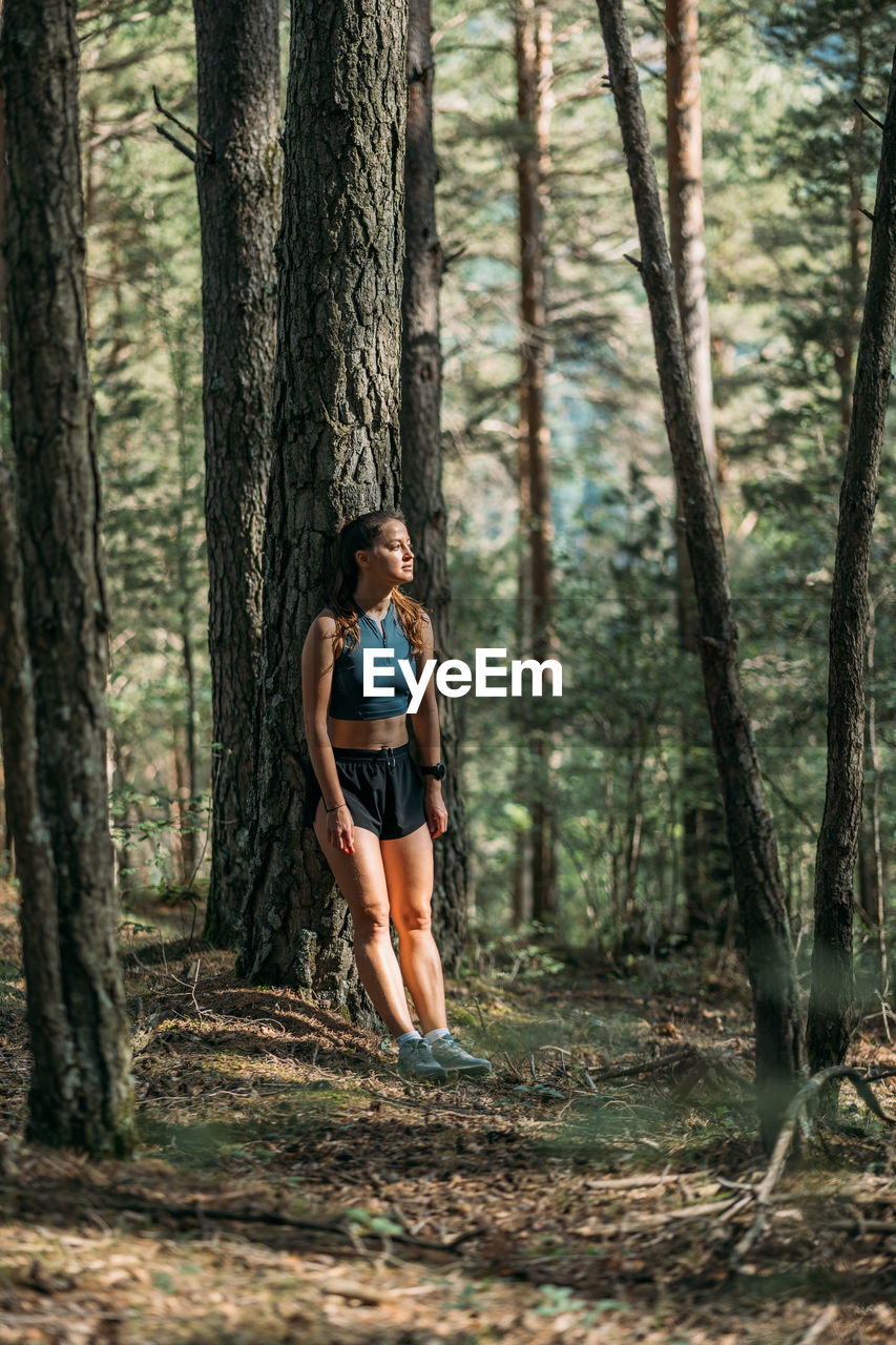 Tired young woman in sportswear leaned on a tree and resting in the forest after working out. 