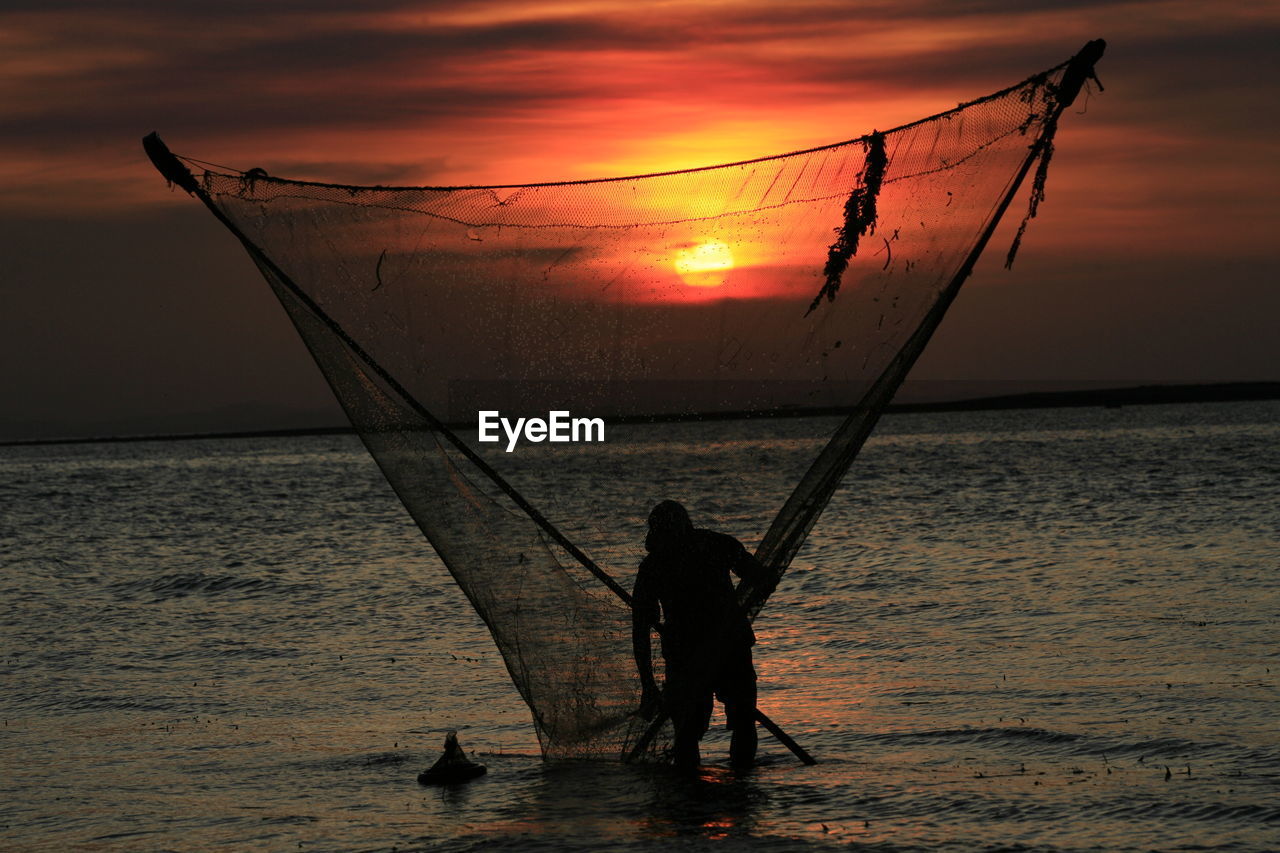 Silhouette fisherman with fishing net in sea against sky during sunset