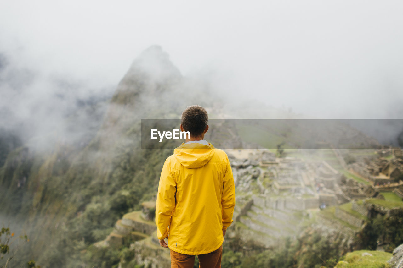 A man in a yellow jacket is standing near ruins of machu picchu, peru