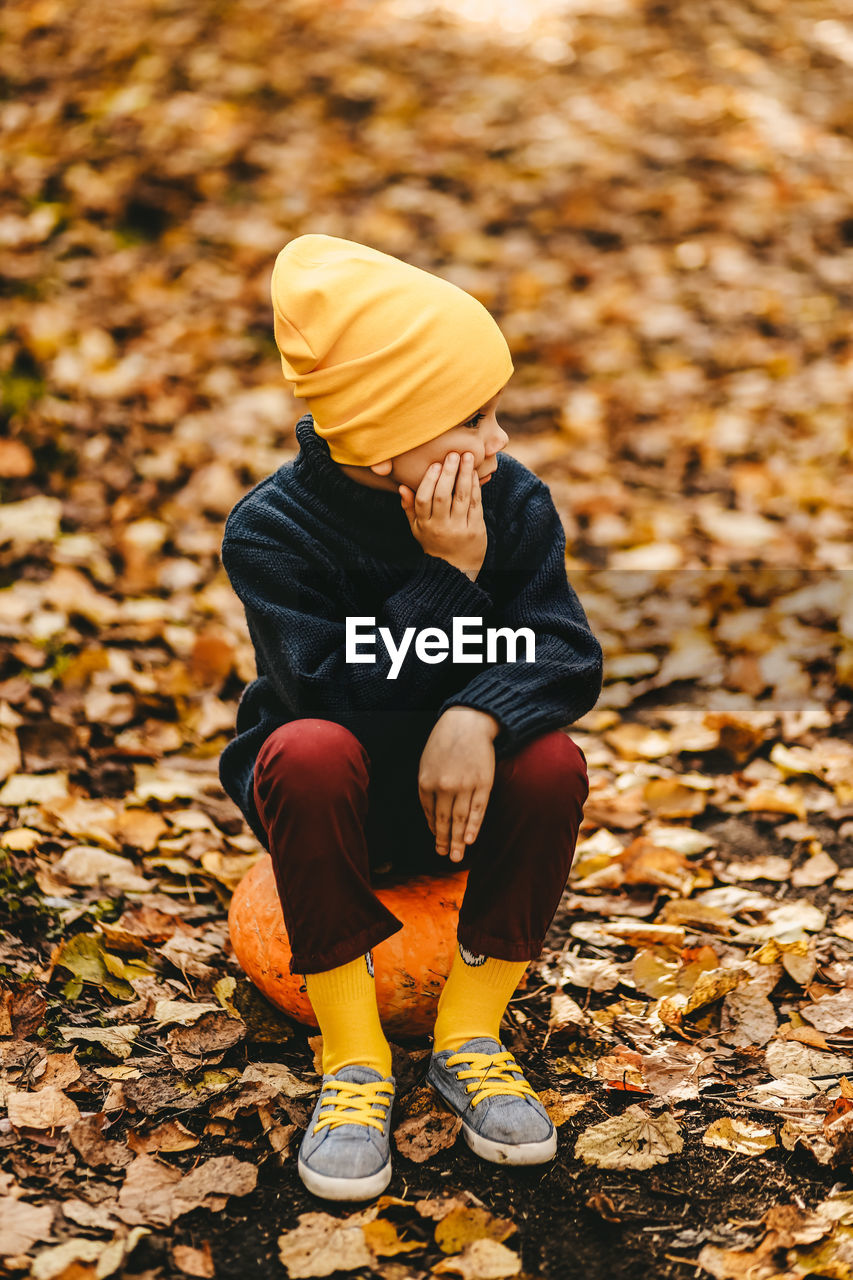 A boy a teen child in a bright yellow hat sits on a pumpkin in an autumn forest in nature outdoors