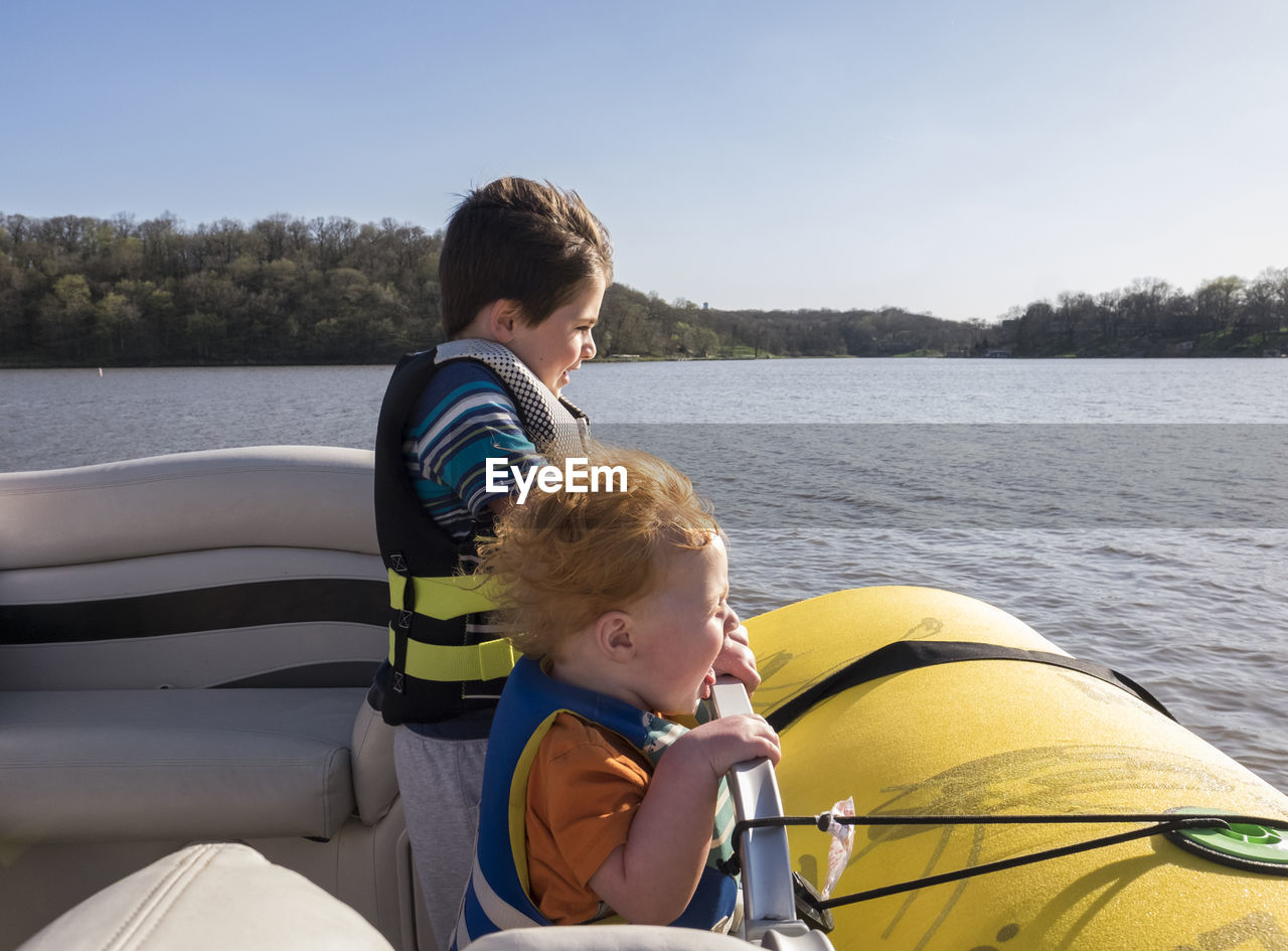 Happy brothers wearing life jackets while standing in boat on lake against clear sky during sunny day
