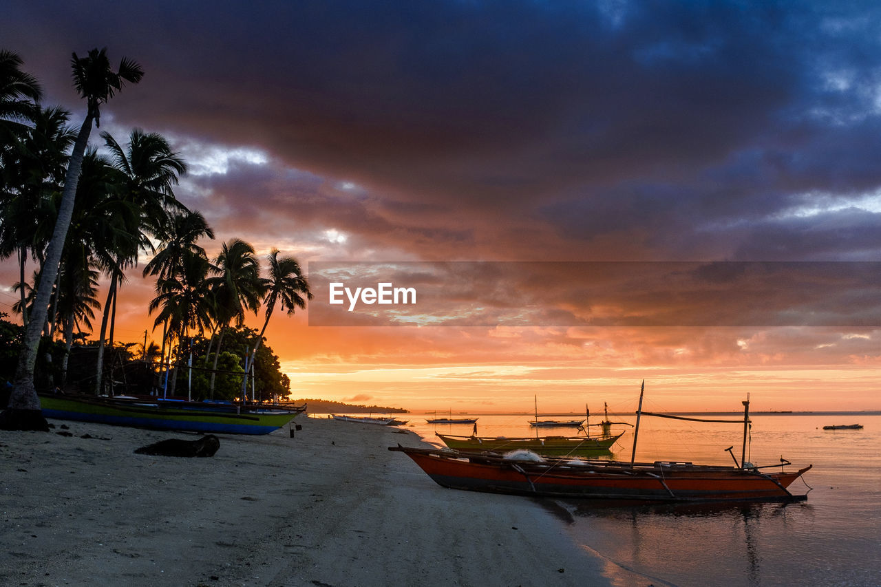 Boats moored on shore against cloudy sky during sunset