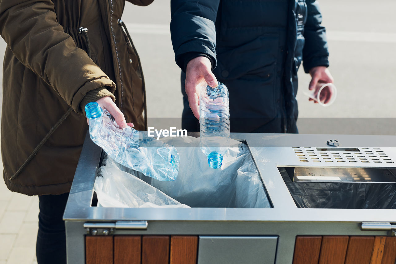 Midsection of people throwing bottles in garbage bin