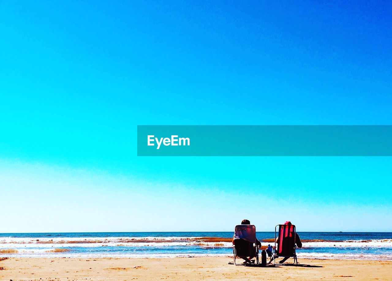 Friends sitting on deck chairs at beach against clear blue sky