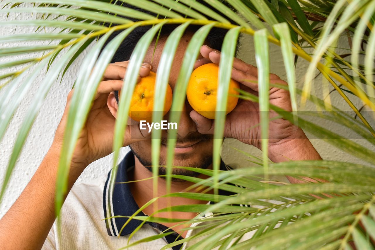 Man covering eyes with orange fruit by plants against wall