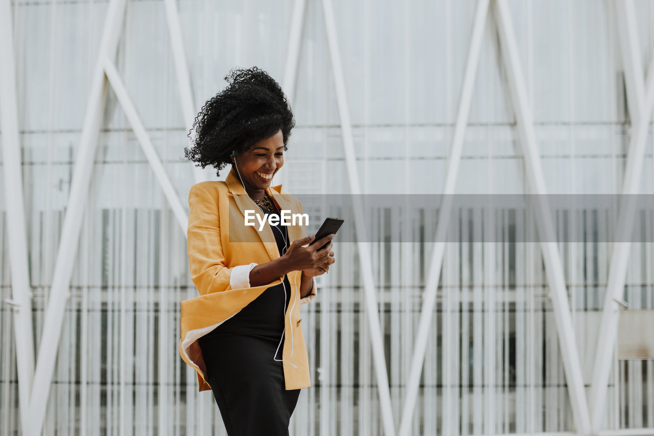 Smiling female entrepreneur using smart phone listening to music through earphones standing against building