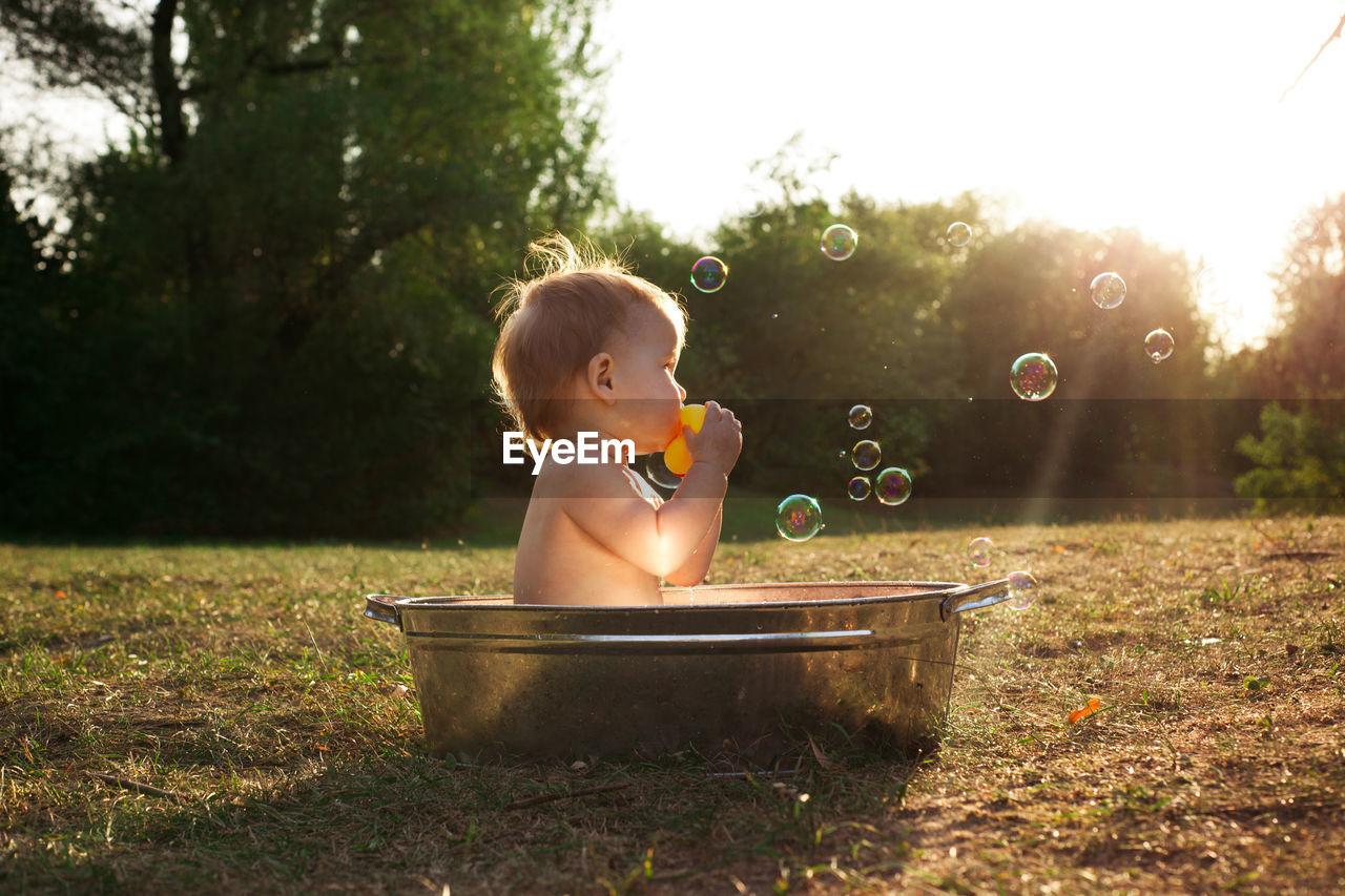 Cute little kid sits in a basin of water in nature and has rays of the setting sun, golden bathe. 
