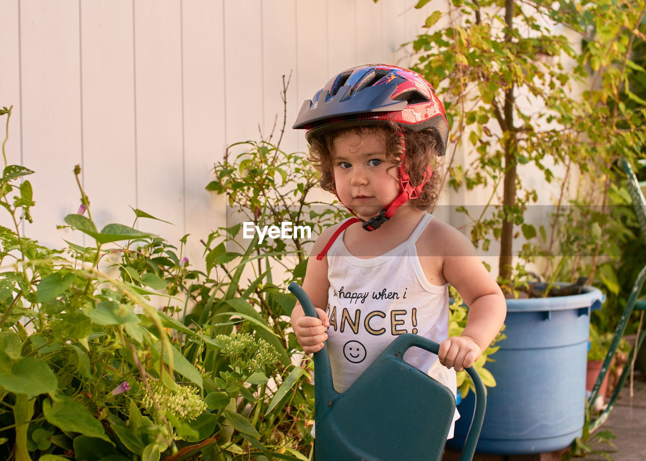 Little girl in a bike helmet is playing in the backyard with plants and watering them