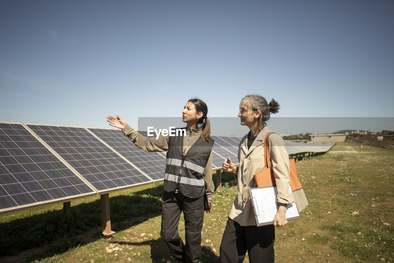 Female engineer showing solar panels to senior entrepreneur holding clipboard while standing at power station