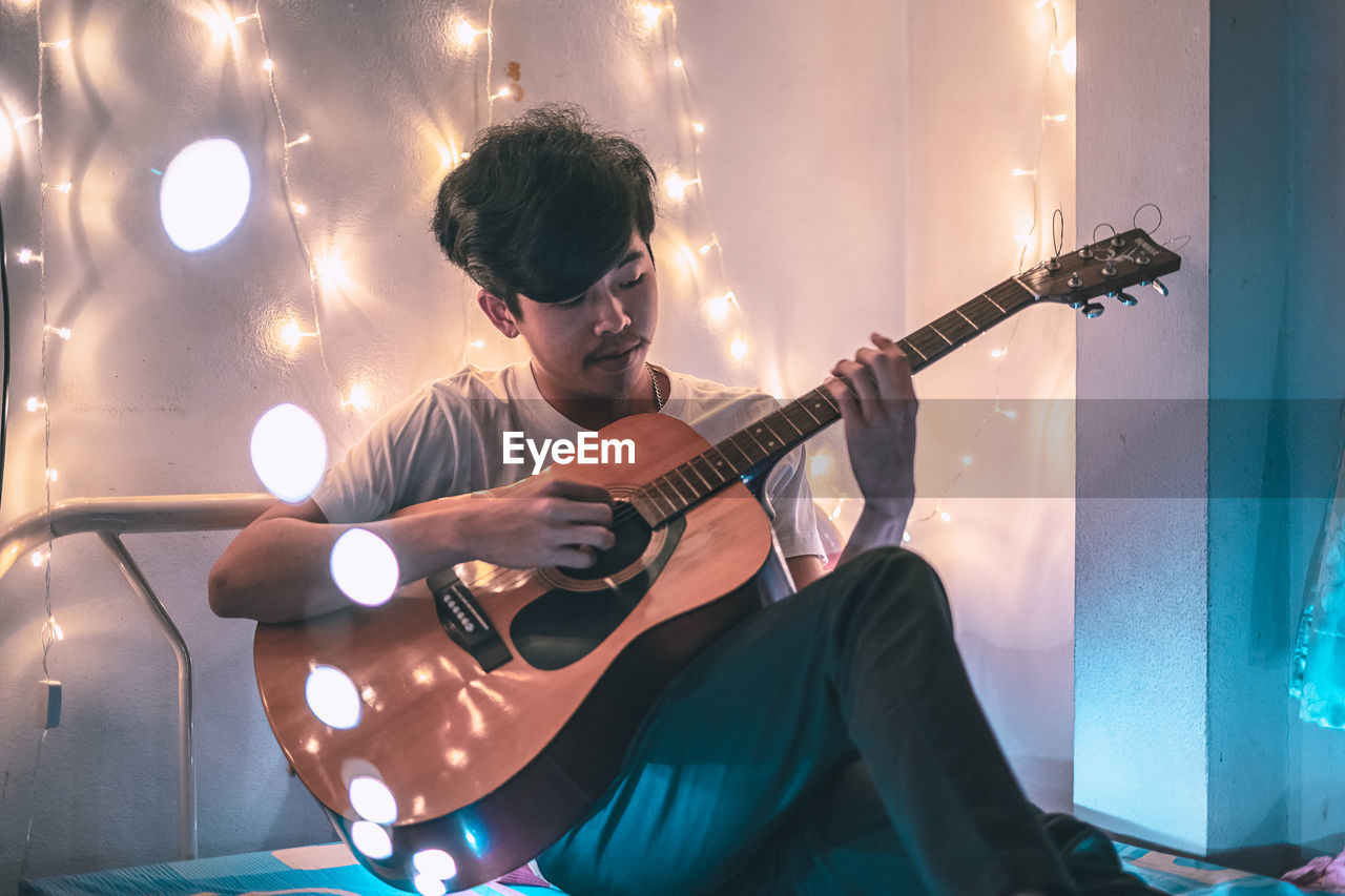 Young man playing guitar while sitting on bed against wall at home