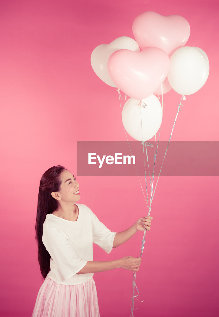 Portrait of happy woman holding heart shape helium balloons while standing against pink background