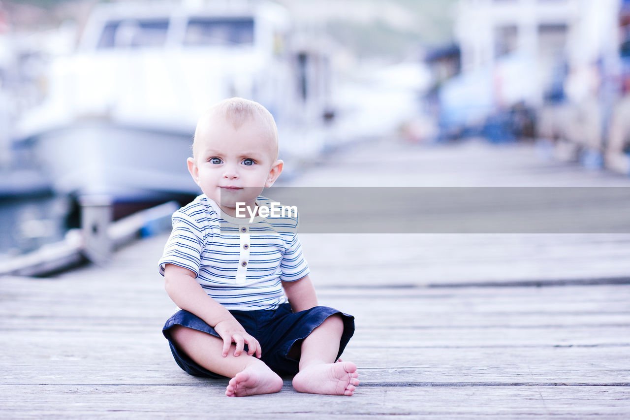 Funny baby boy wearing striped shirt sitting on wooden pier outdoors. looking at camera