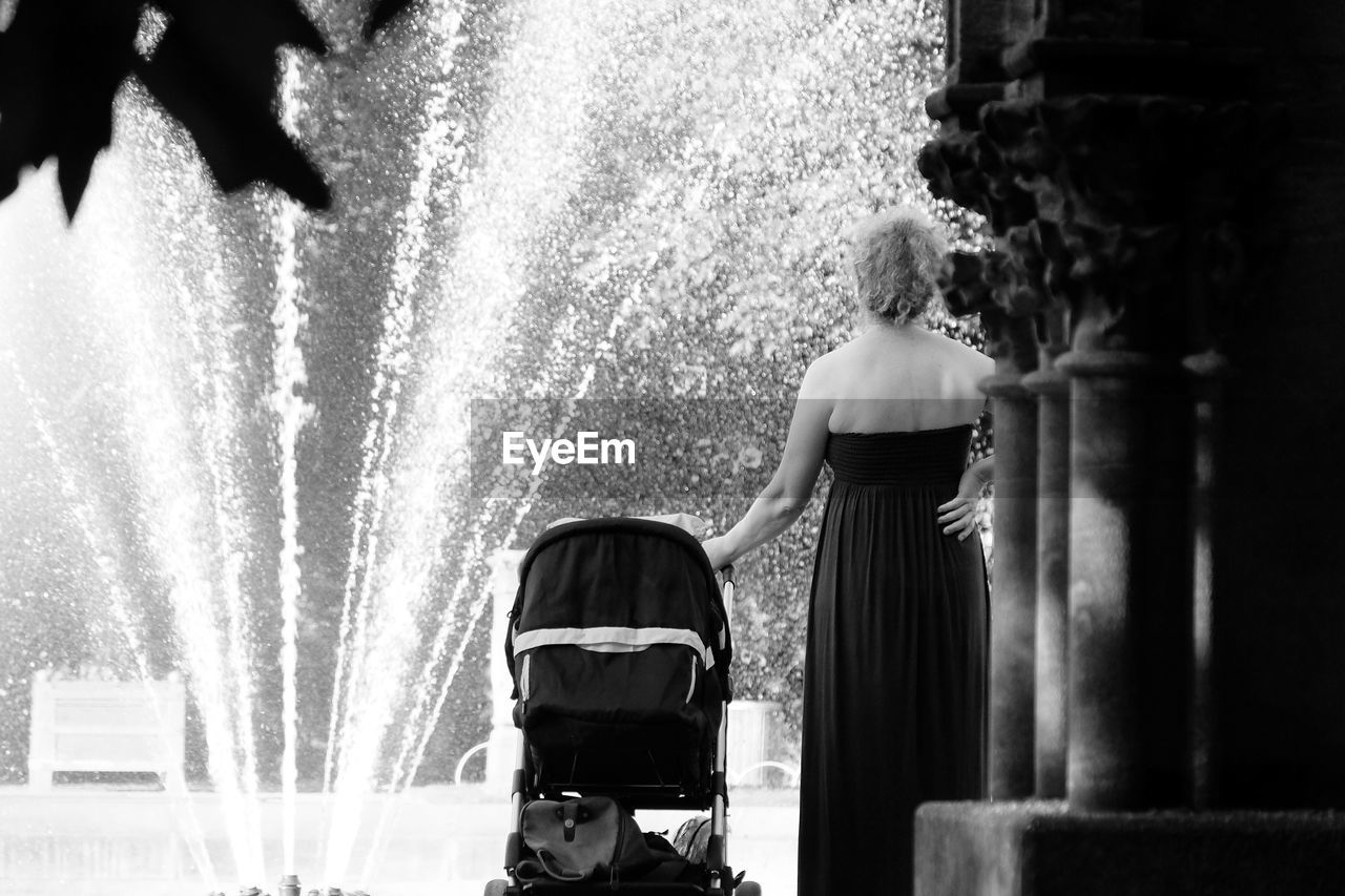 Rear view of woman standing with baby carriage against fountain