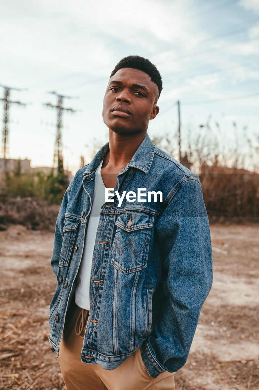 Young serious african american male with modern haircut in denim jacket looking at camera under cloudy sky in fall