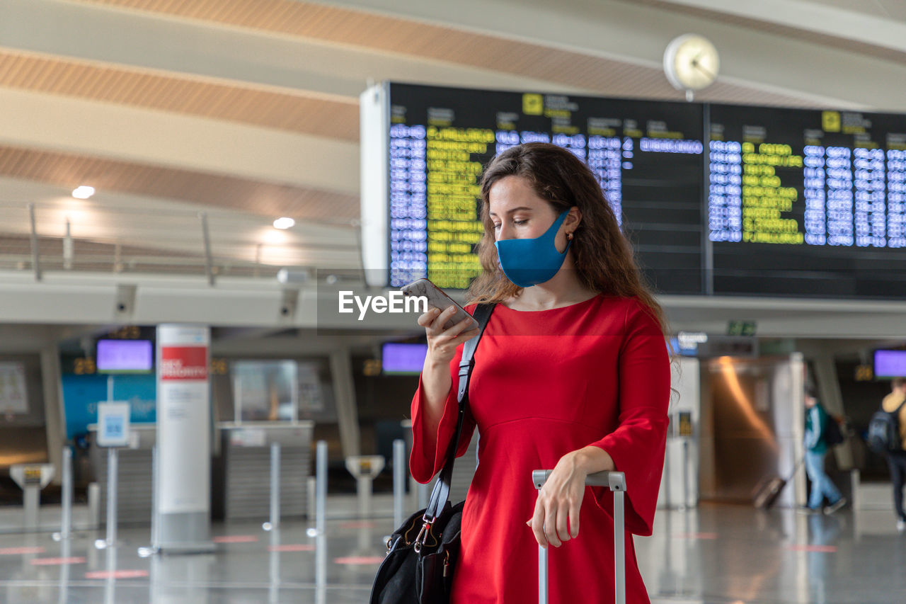 Serious female traveler in mask standing with suitcase on background of departure board and browsing cellphone while waiting for flight
