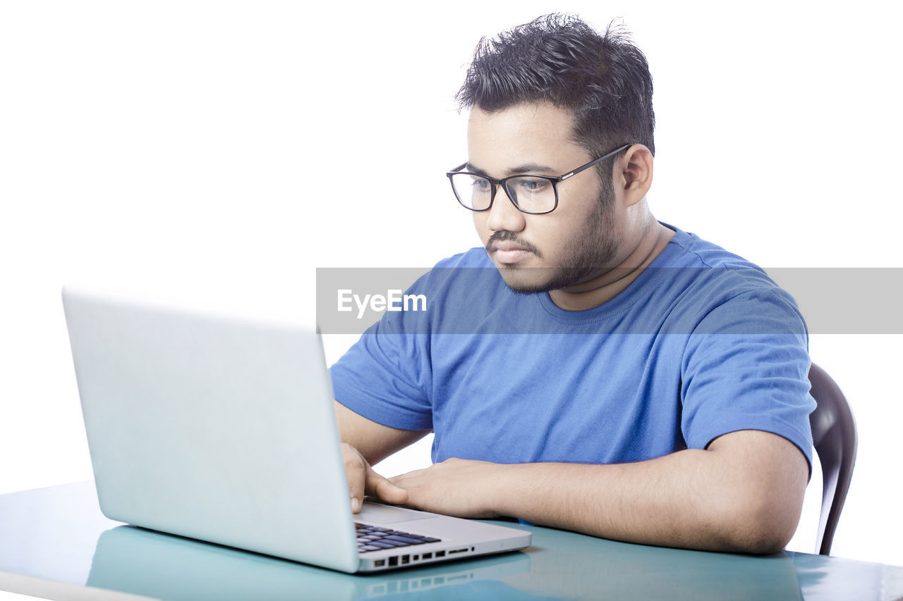 technology, computer, wireless technology, laptop, eyeglasses, using laptop, one person, glasses, communication, men, adult, internet, writing, computer network, business, casual clothing, indoors, looking, person, sitting, table, young adult, typing, e-mail, working, conversation, businessman, computer equipment, concentration, office, furniture, relaxation, desk, front view, portrait, portability, business finance and industry, lifestyles, copy space, student, using computer, learning, white background, education, occupation