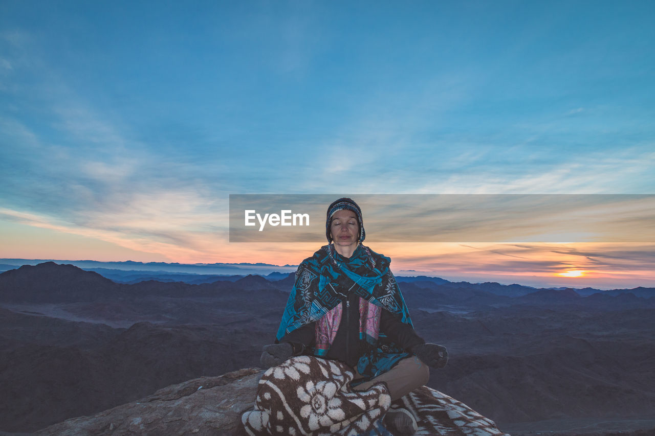 Woman meditating while sitting on rock against sky during sunset