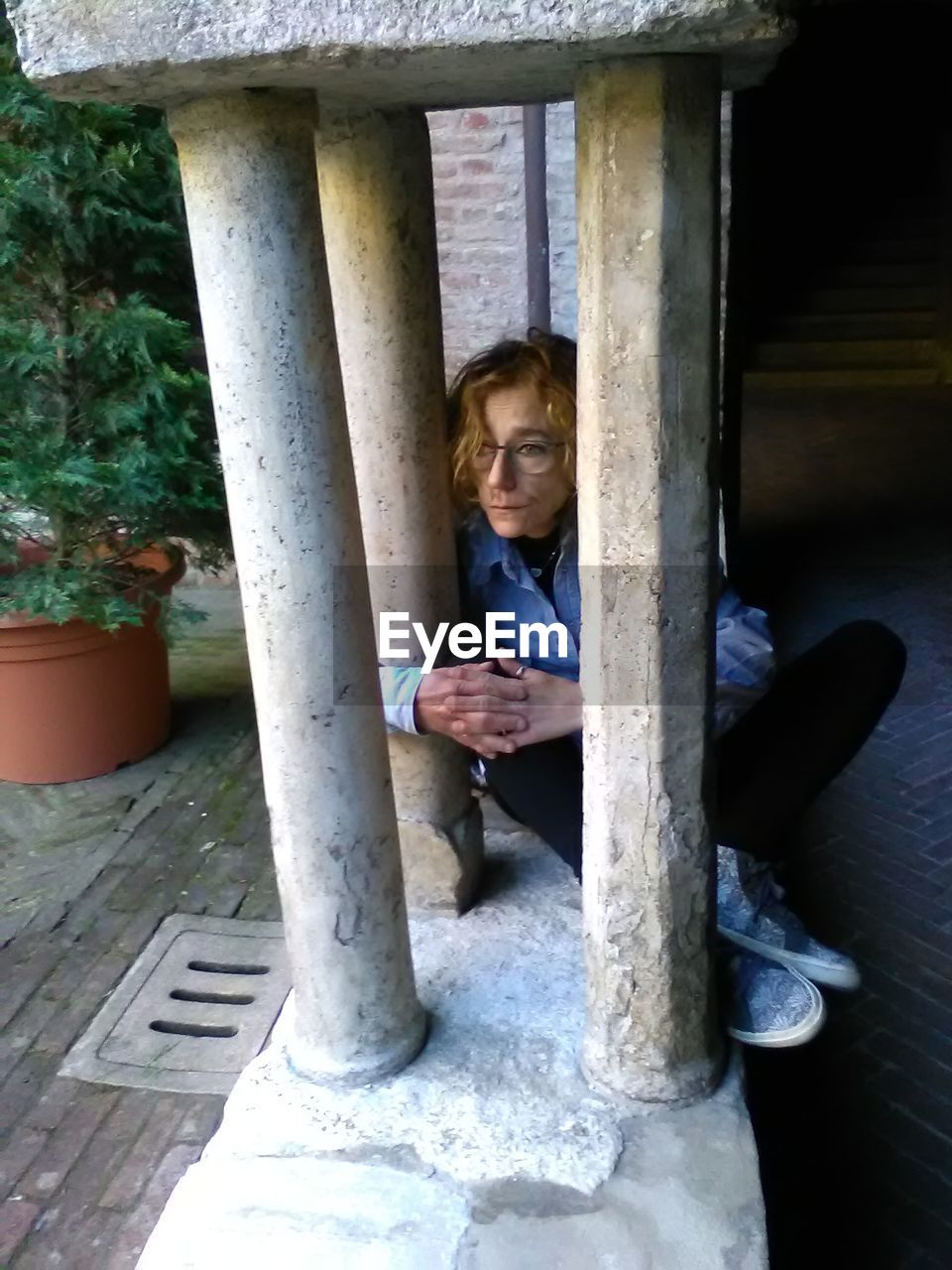 Full length of thoughtful mid adult woman looking through columns while sitting on retaining wall