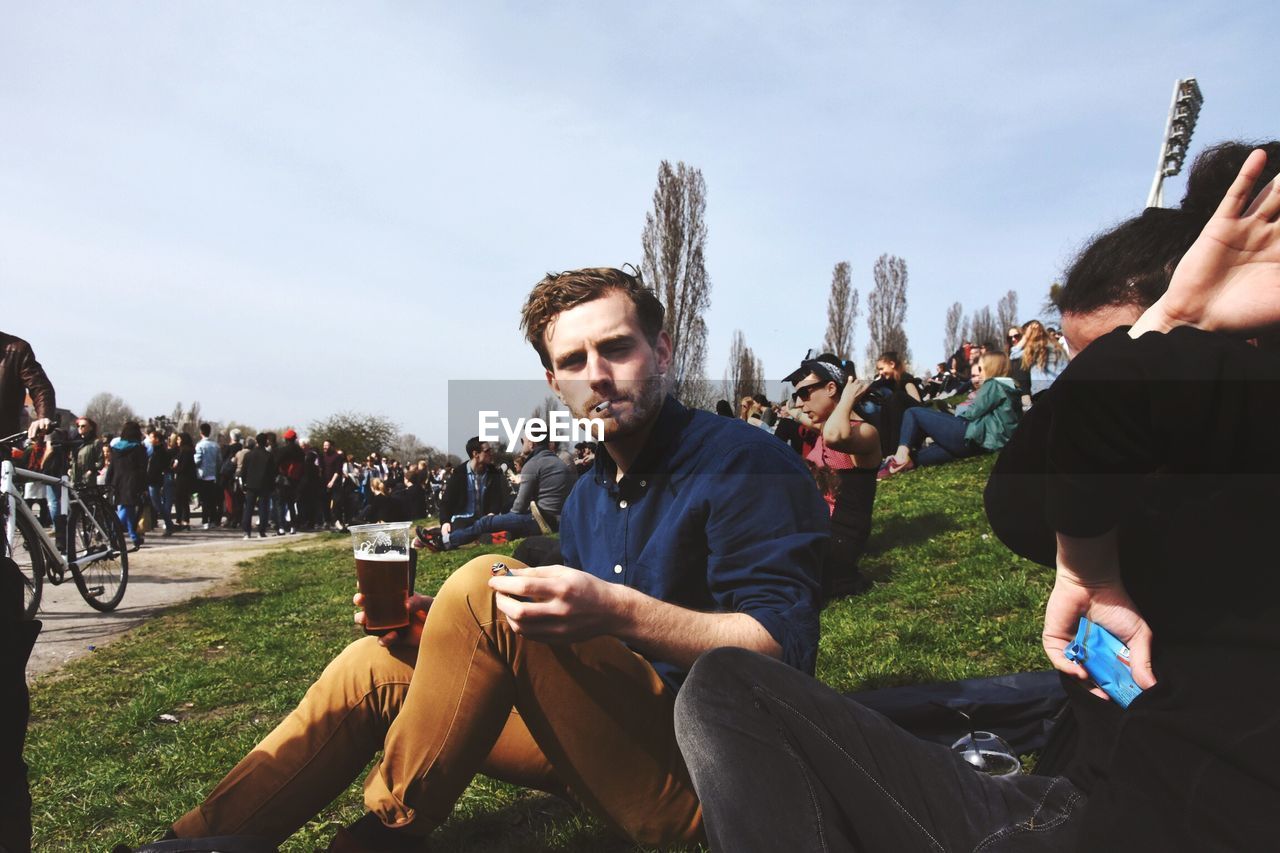 Portrait of man smoking cigarette while sitting with people on grassy field