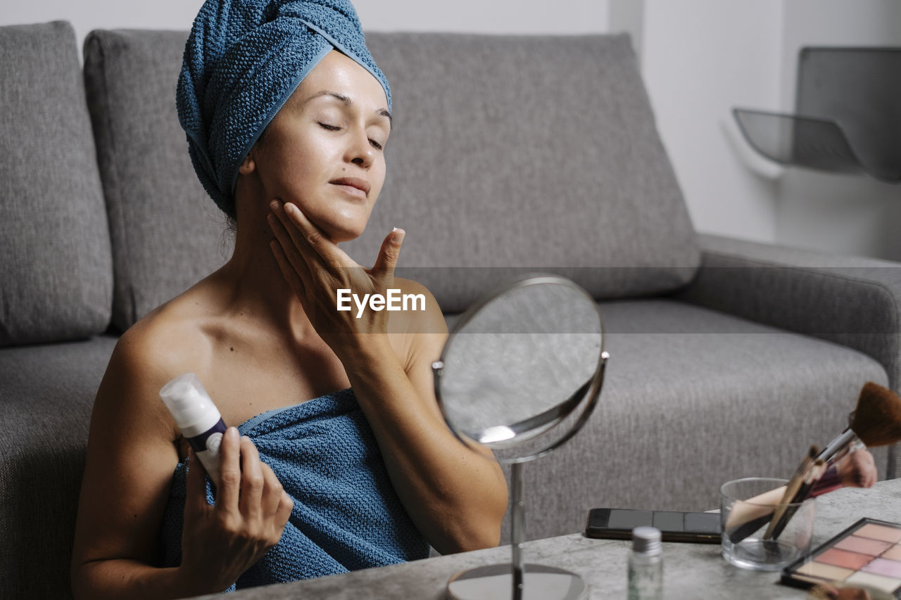 Charming young female with bare shoulders and towel on head smearing facial skincare cream on face during makeup procedure at home