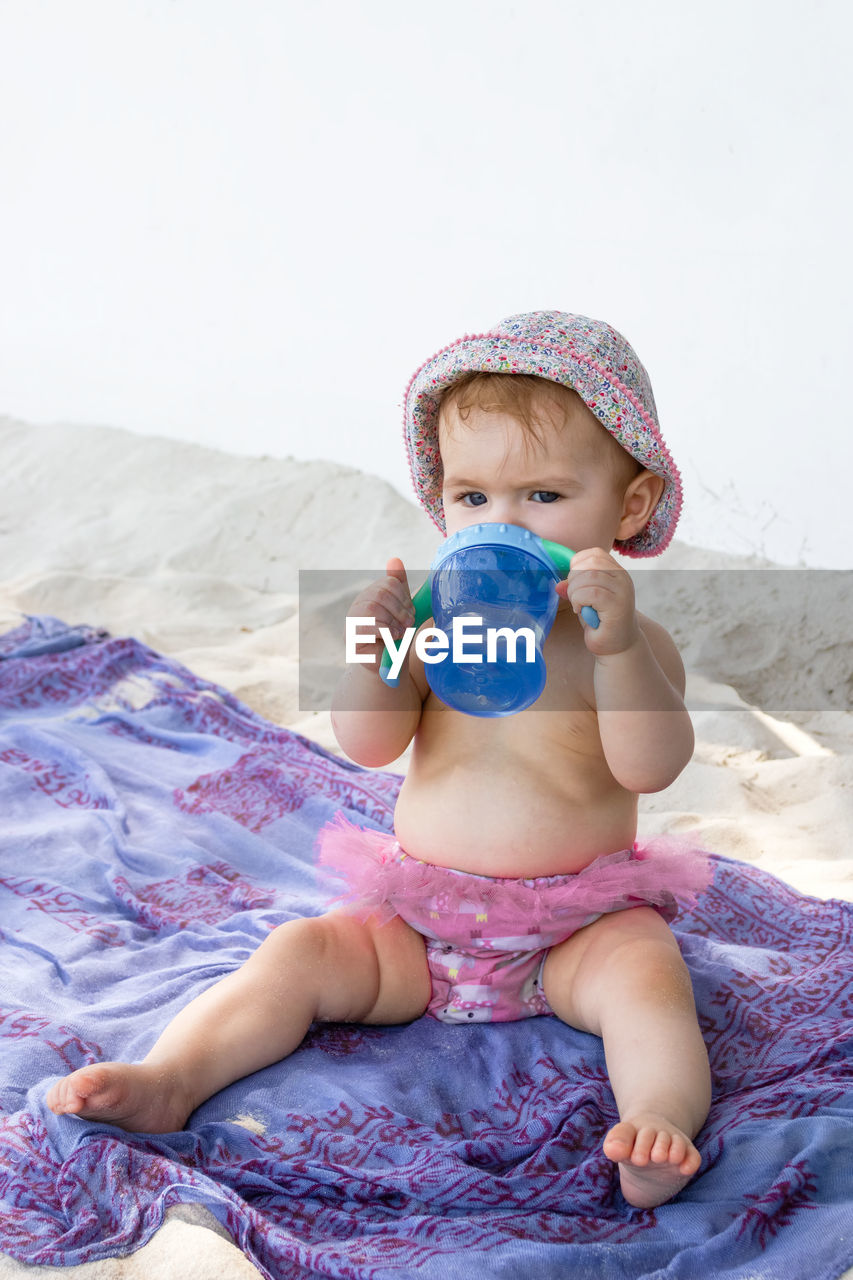 Little baby girl  sitting on plaid at beach and drinking water from bottle.