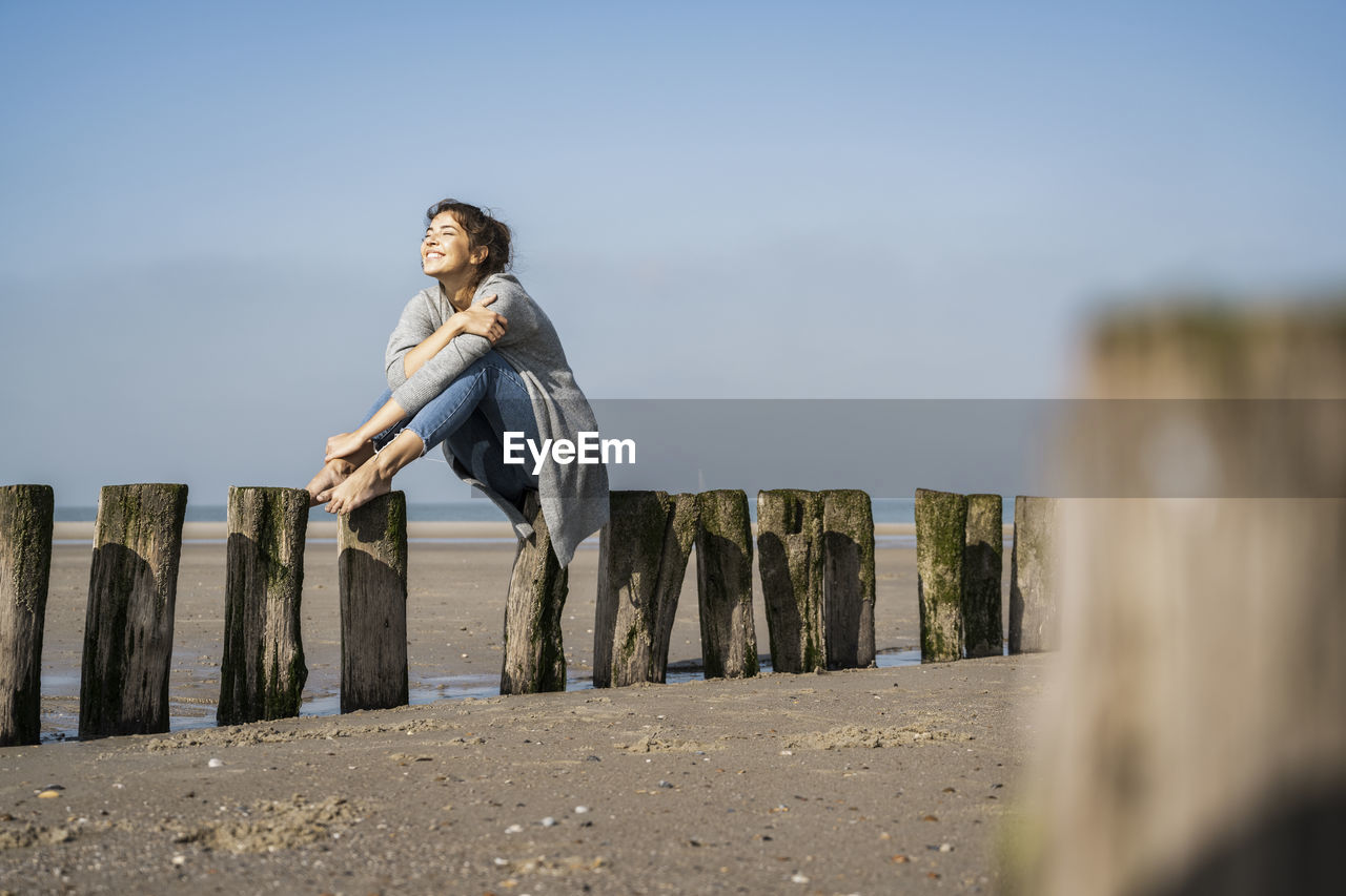 Smiling young woman sitting on wooden posts at beach against sky