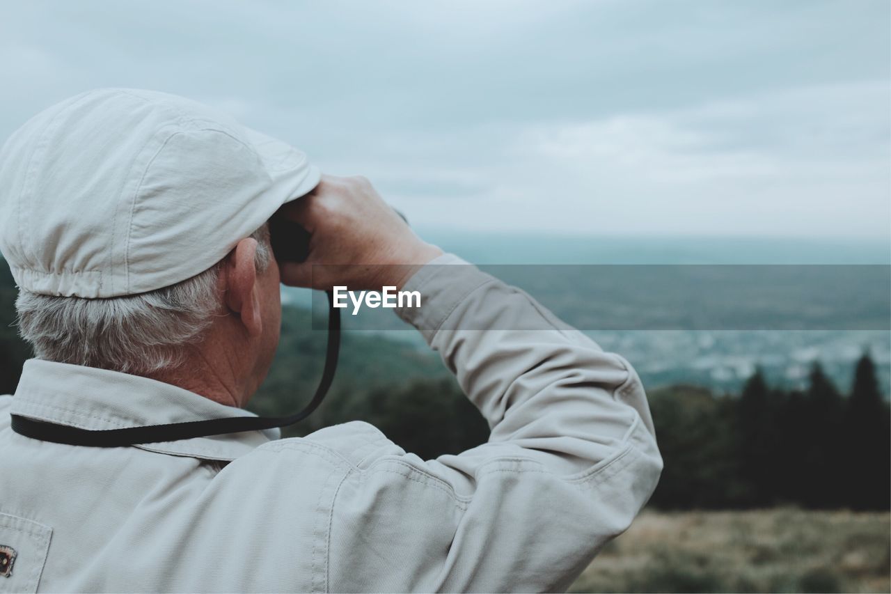 Rear view of senior man looking through binoculars while standing on mountain against cloudy sky
