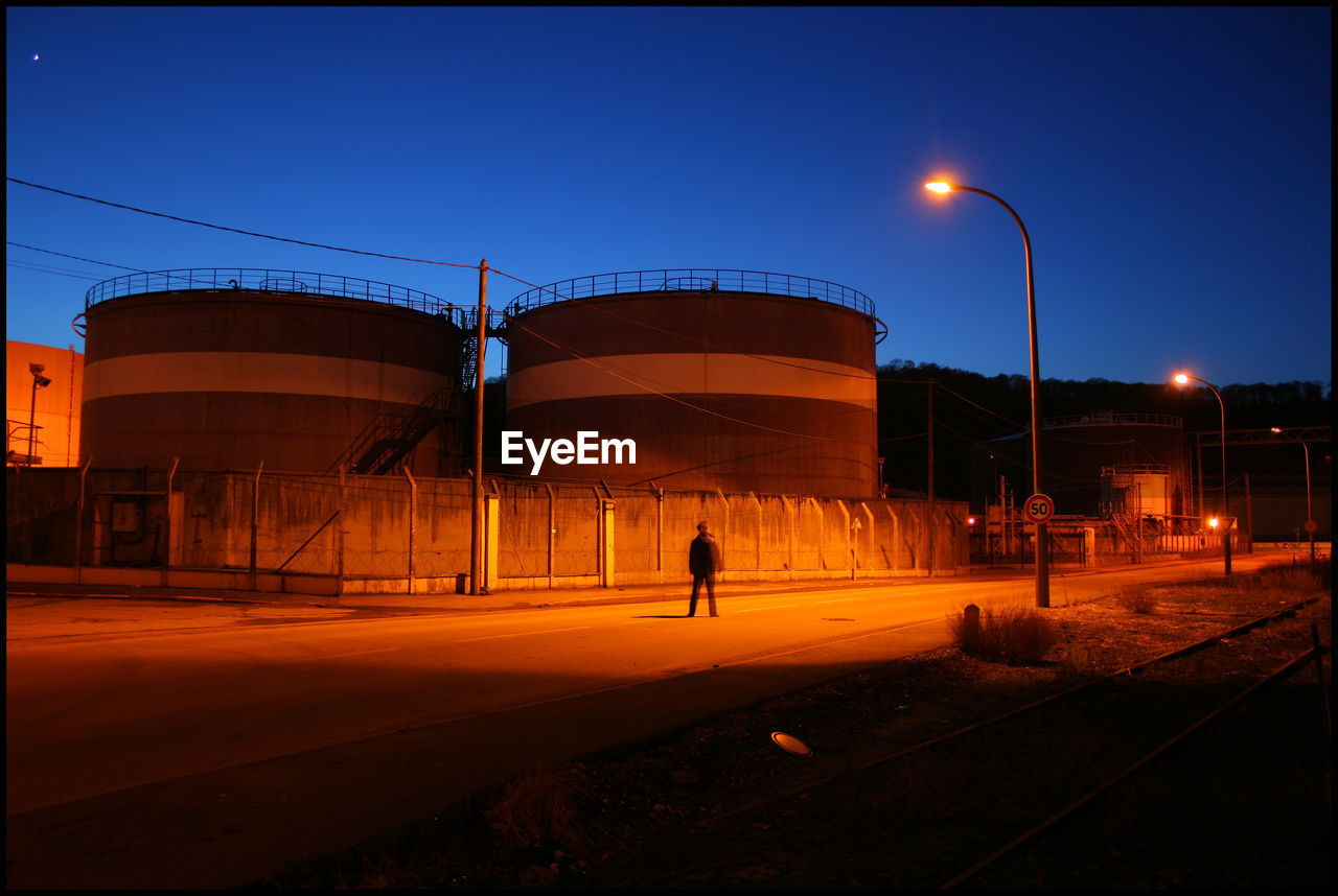 Person walking on road by industry against blue sky at night