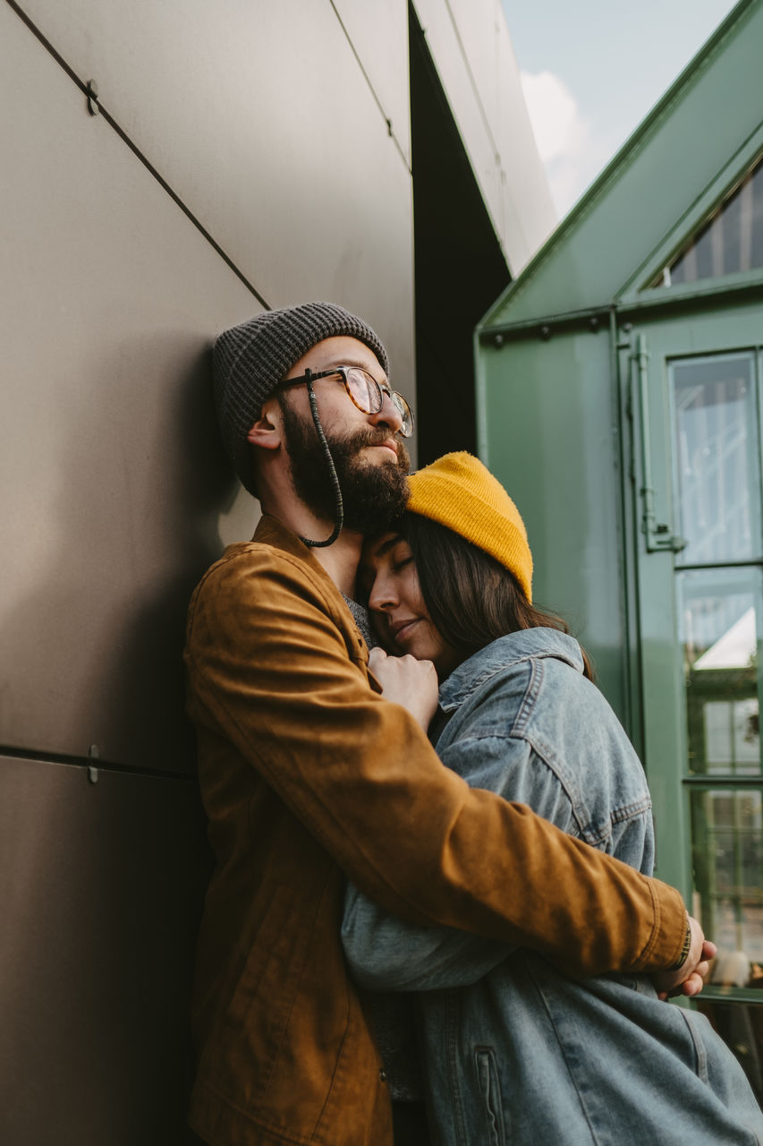 Side view of young stylish couple hugging gently while standing on wooden terrace near house