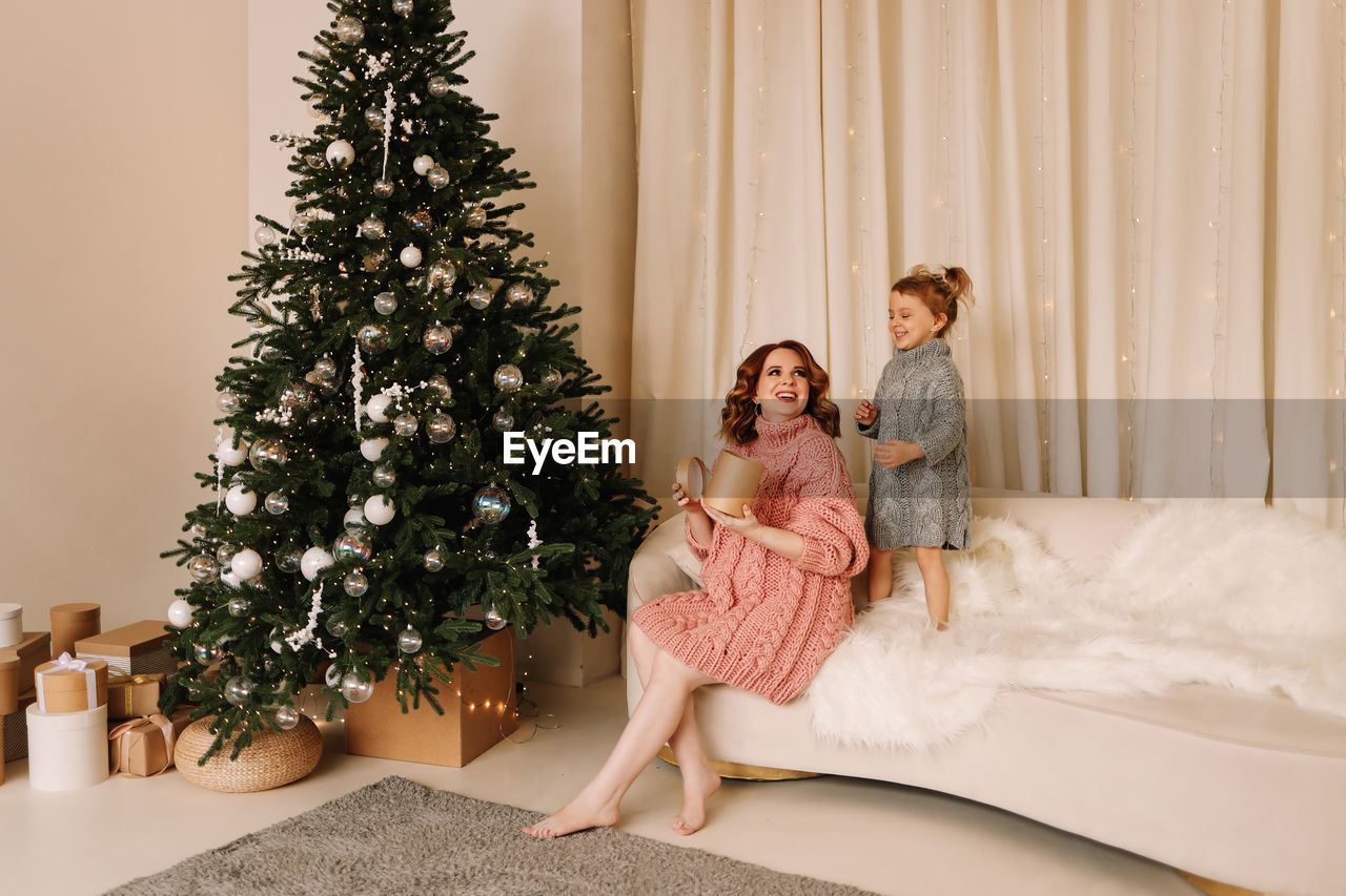 women, holiday, celebration, christmas, christmas tree, tree, adult, emotion, happiness, female, togetherness, plant, two people, indoors, smiling, domestic room, full length, decoration, positive emotion, men, lifestyles, domestic life, nature, interior design, gift, family, love, sitting, home interior, child, young adult, childhood, cheerful, living room, tradition, bonding, anticipation, enjoyment, dress, christmas decoration, clothing, furniture, event, brown hair, person, standing, parent
