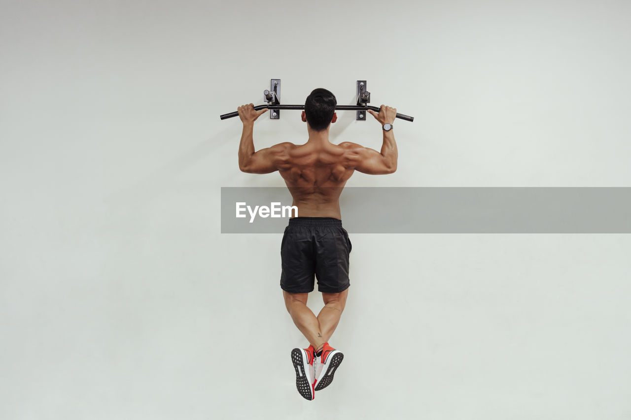 Mid adult sportsman doing chin-ups with exercise equipment on white wall