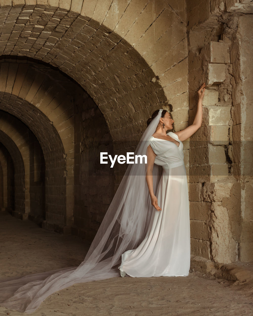 A sad elegant lady in white bride dress with a train languishes in the gloomy old castle