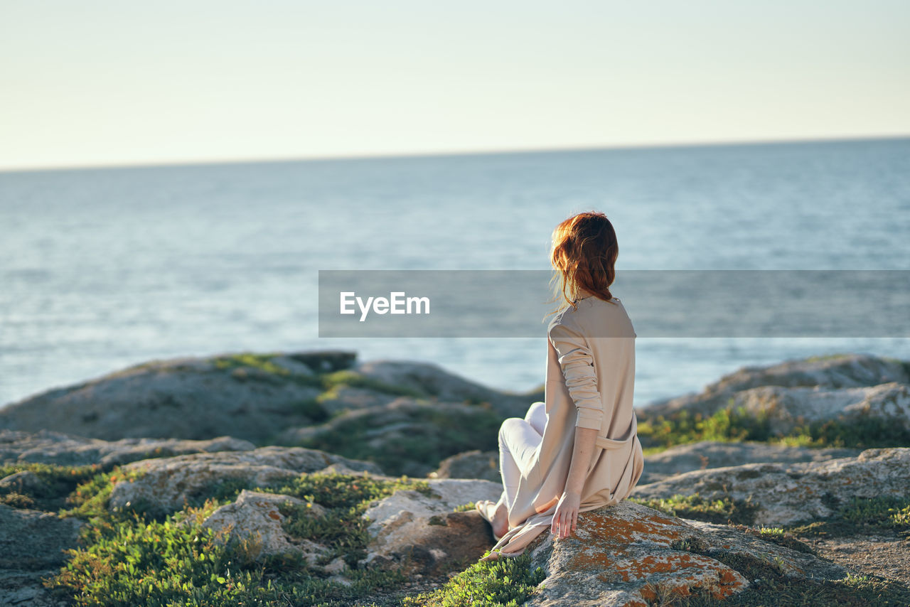 REAR VIEW OF WOMAN SITTING ON ROCK LOOKING AT SEA