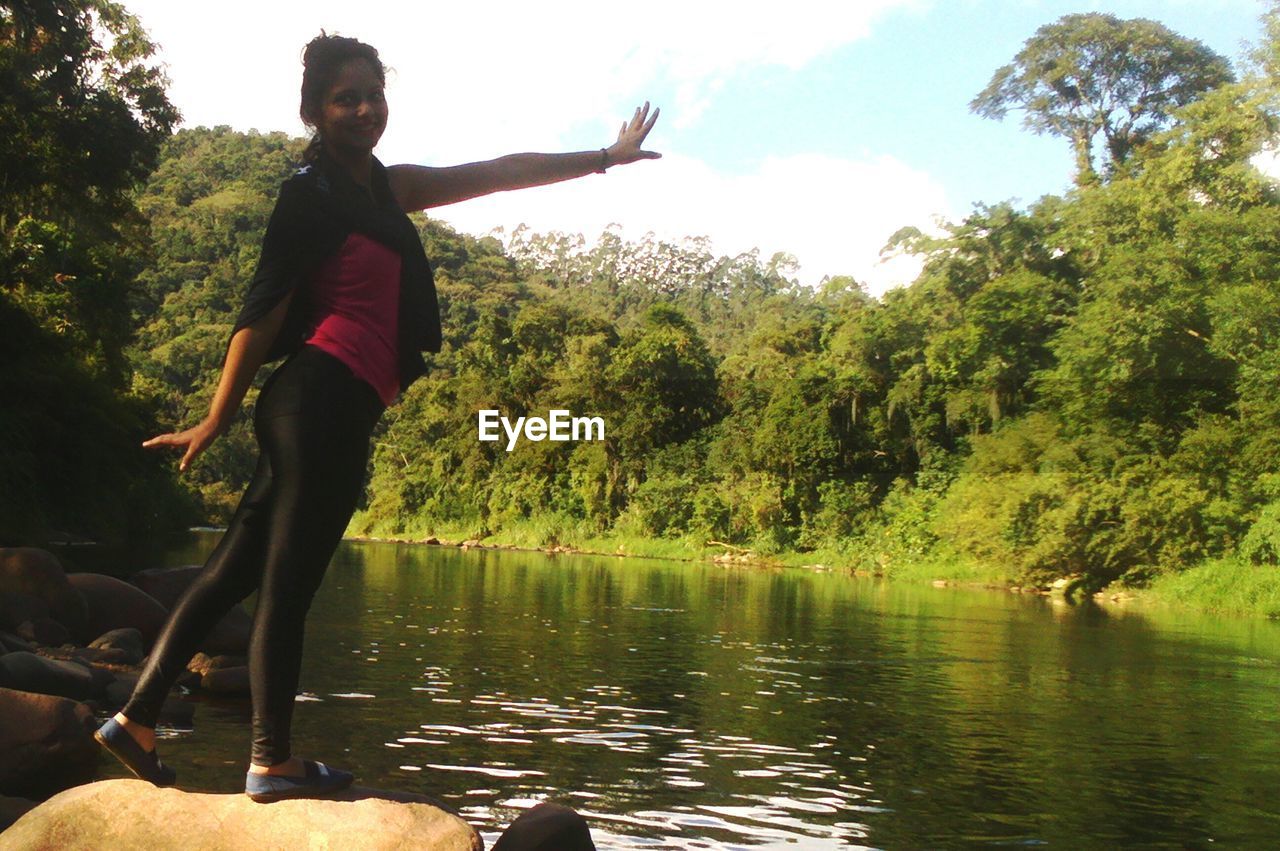 YOUNG WOMAN JUMPING BY LAKE AGAINST TREES