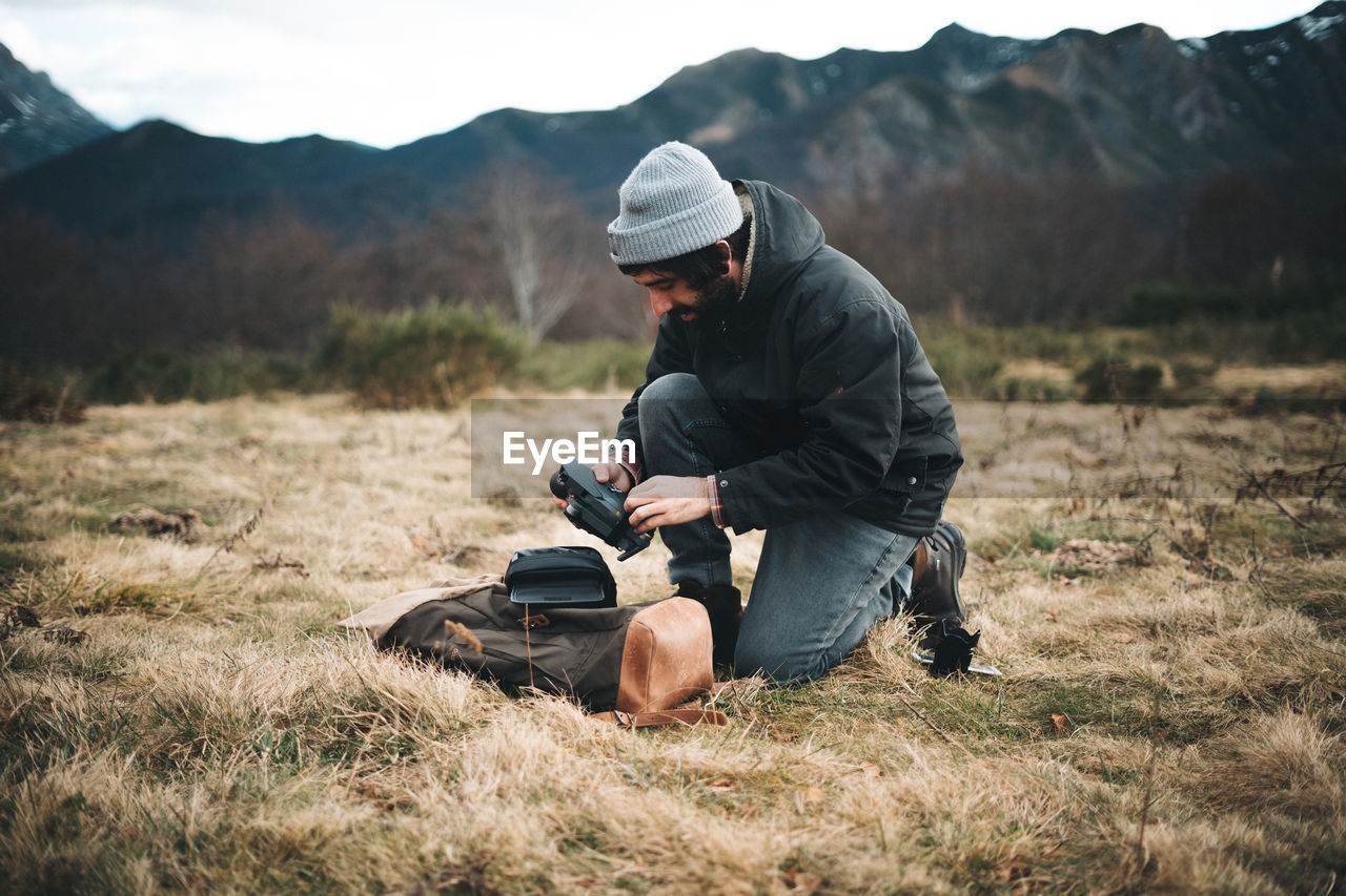 Side view of man in hat and warm jacket taking accessories from camera in meadow with dry grass